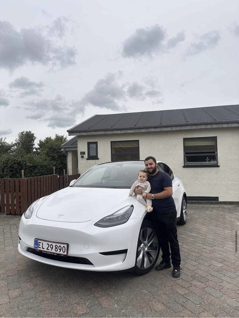 Mand og baby foran hvid bil ved hus